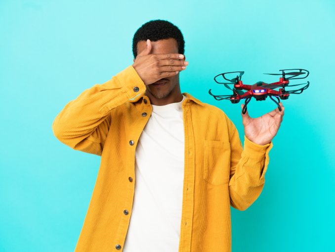 African American handsome man holding a drone over isolated blue background covering eyes by hands. Do not want to see something